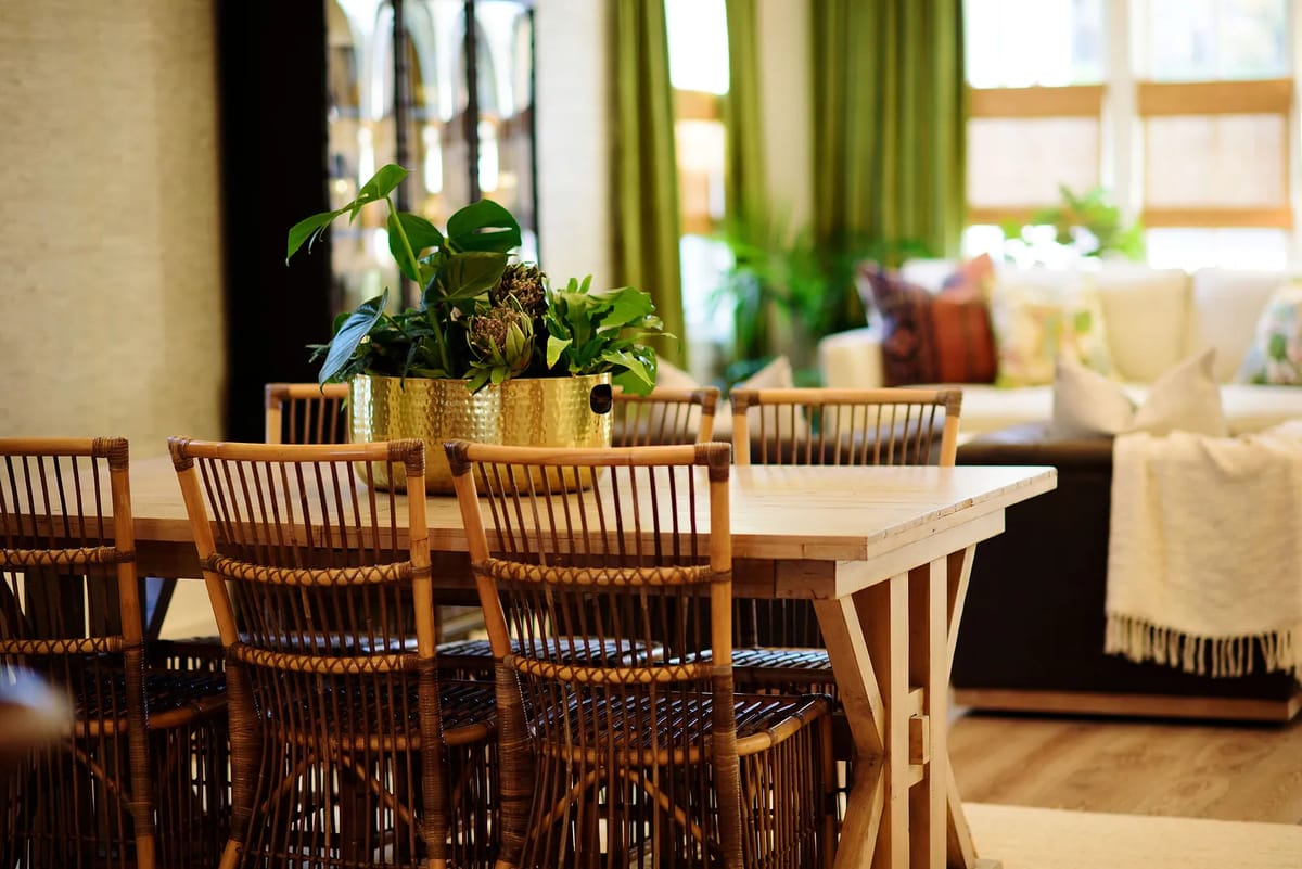 Dining room with wooden table, rattan chairs, planter, and large windows with green curtains.