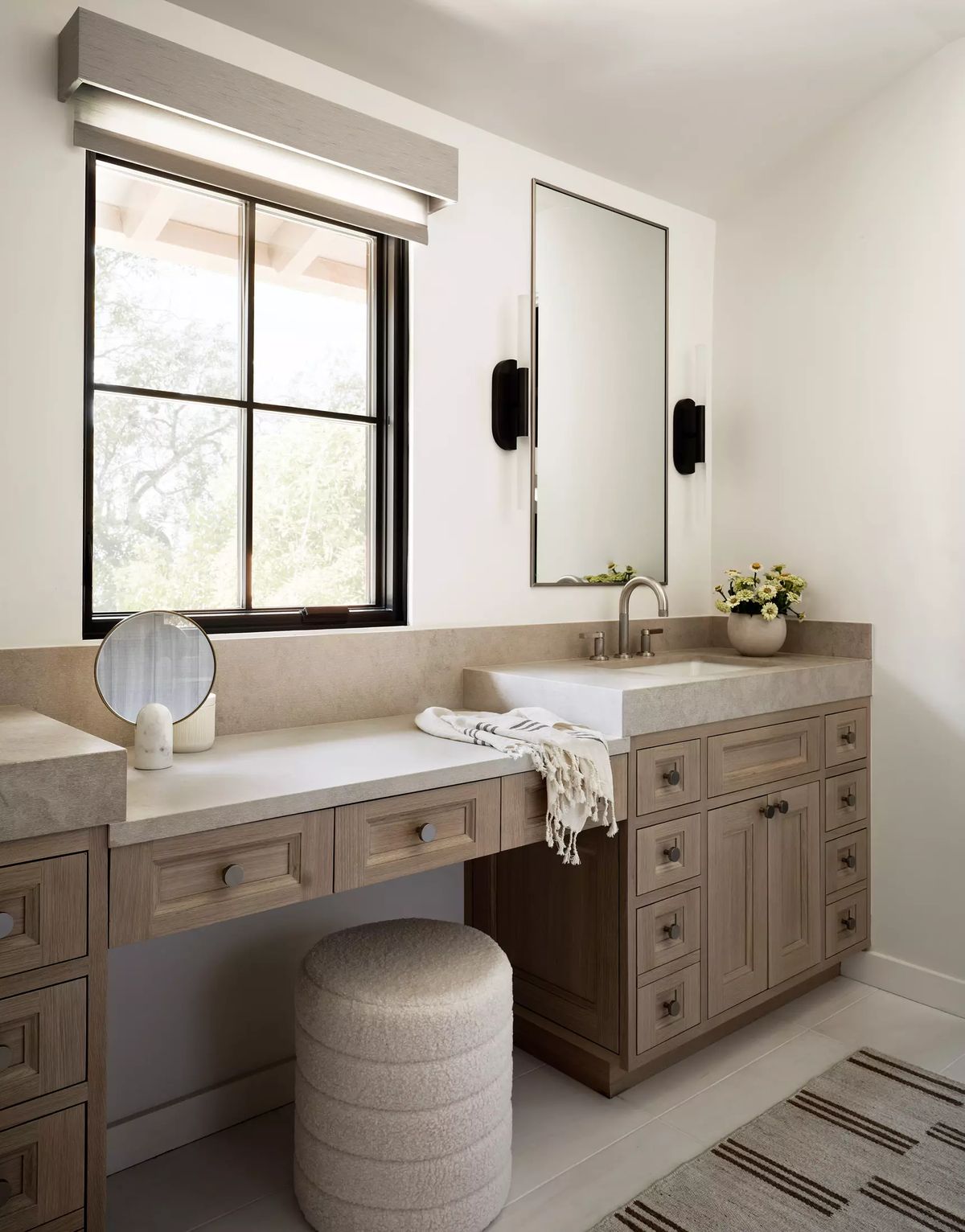 Bathroom with wooden vanity, white countertop, round mirror, circular stool, and patterned rug with natural light.