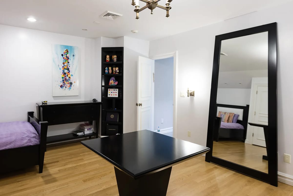 Bedroom with black platform bed, purple duvet, black desk, round table, large mirror, and colorful wall art.