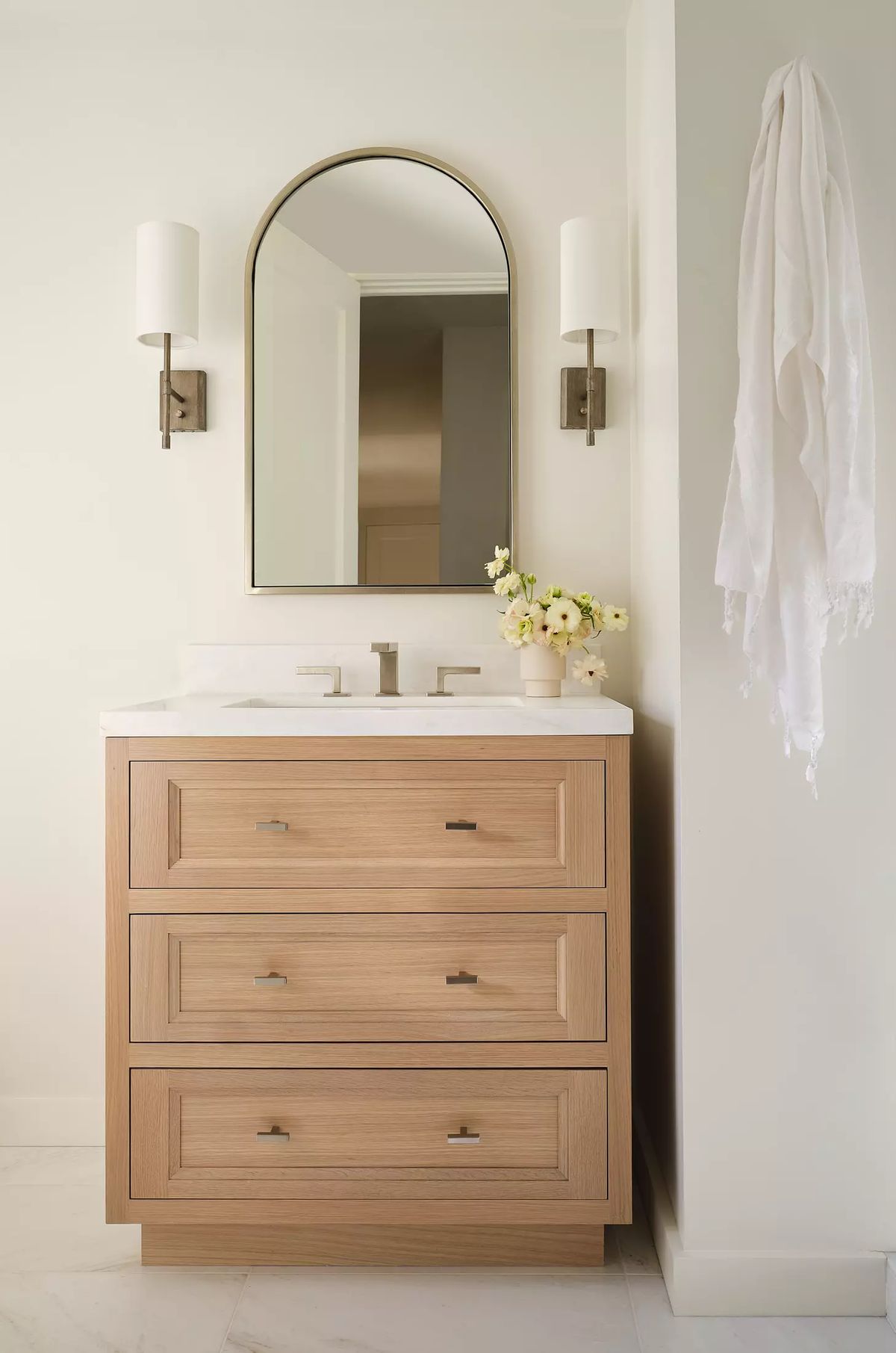 Bathroom with wooden vanity, white countertop, arched mirror, wall sconces, and small vase of flowers.
