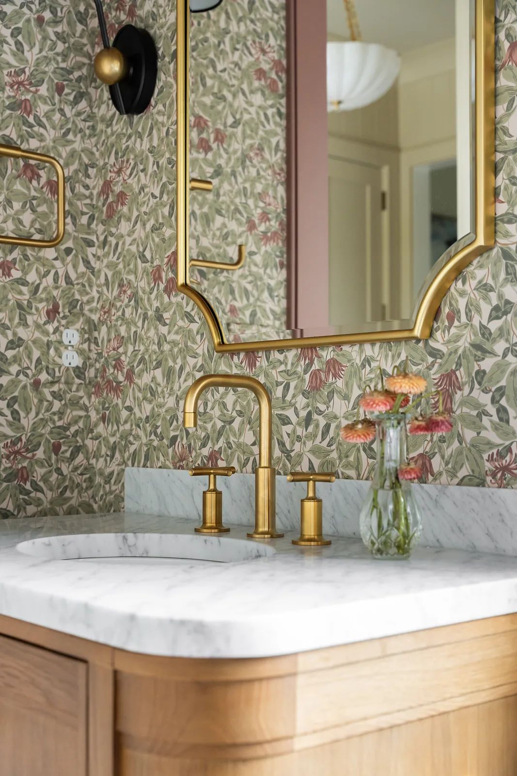 Bathroom with marble countertop, gold faucet, mirror, and floral wallpaper.
