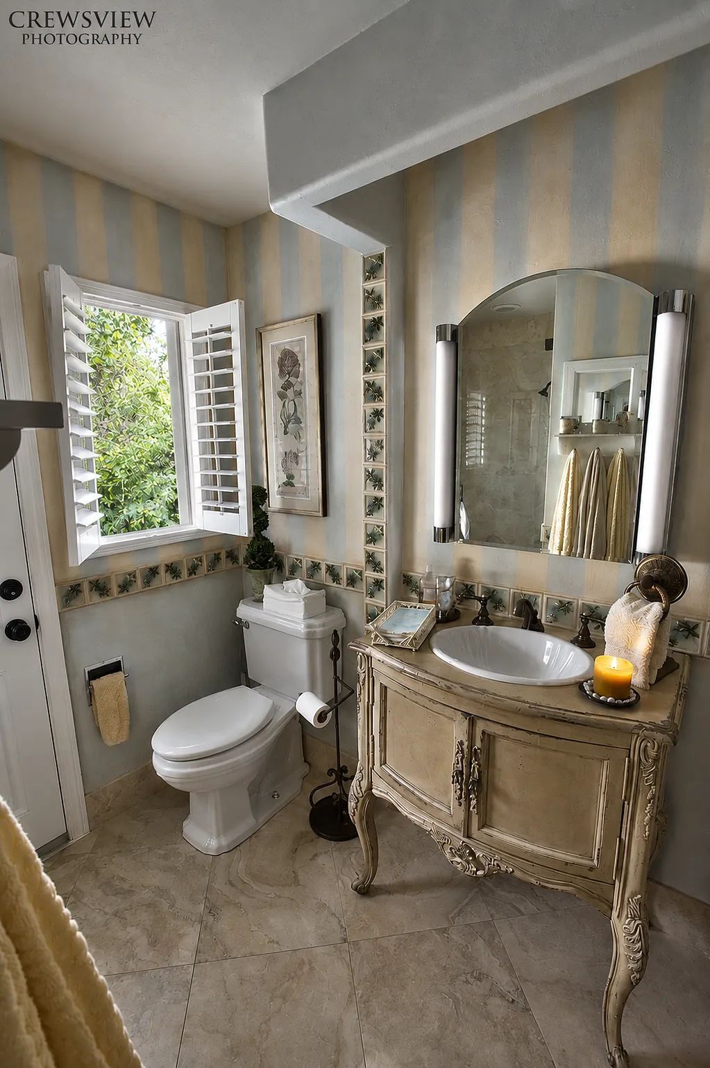 Bathroom with white toilet, vintage sink cabinet, oval mirror, beige wall stripes, and decorative floral border.