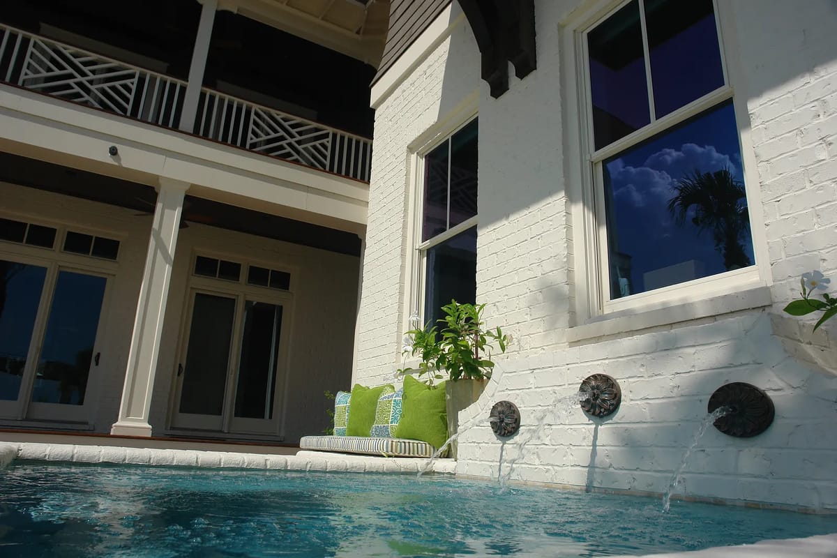 Patio with swimming pool, water fountains, green pillows, and white brick wall with wooden beams.