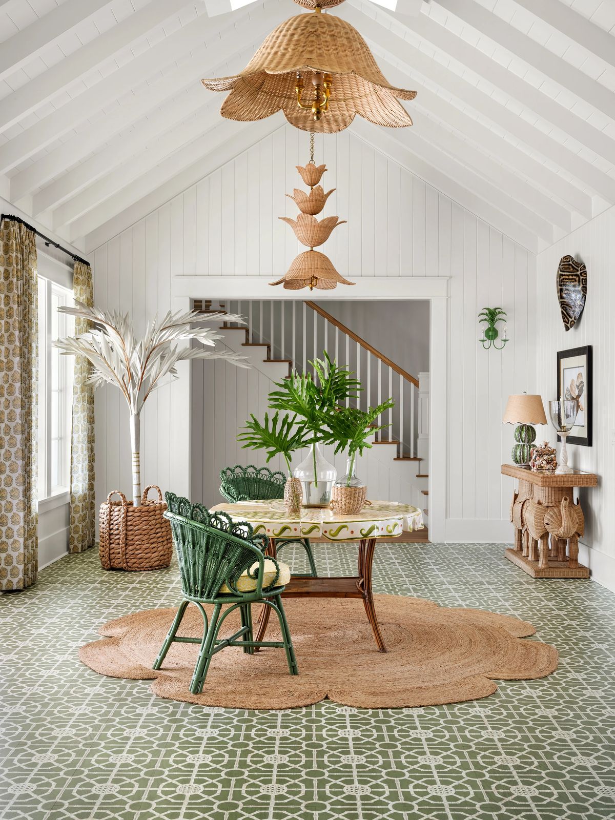 Dining room with round table, green wicker chairs, area rug, potted plants, and decorative wall features.