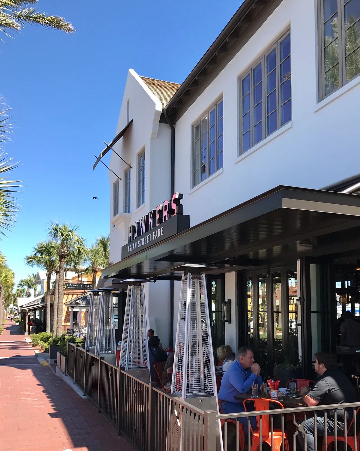 Exterior front view of 'Hawkers Asia Street Fare' with seating area, palm trees, and large windows.