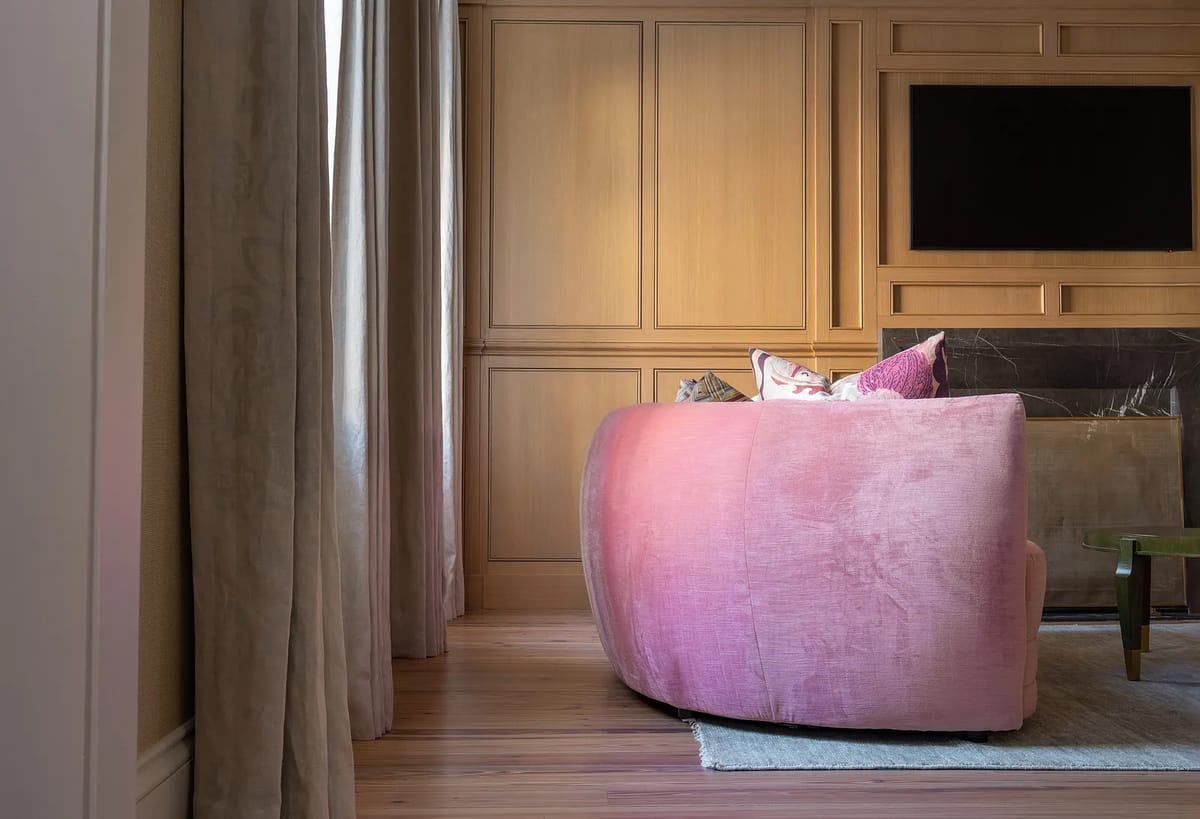 Living room with curved pink sofa, green side table, cushions, wooden panels, and large windows with drapes.