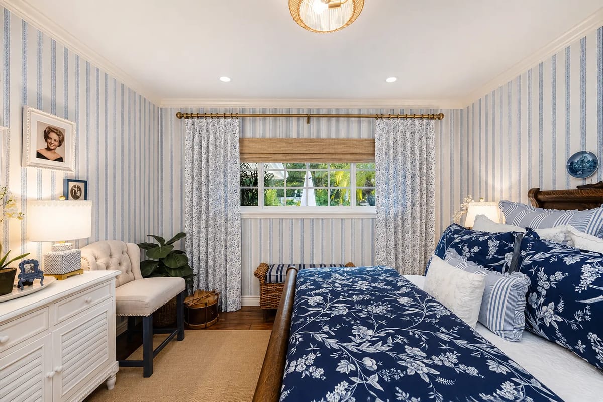 Bedroom with floral bedding, side table, chair, and window with striped curtains.