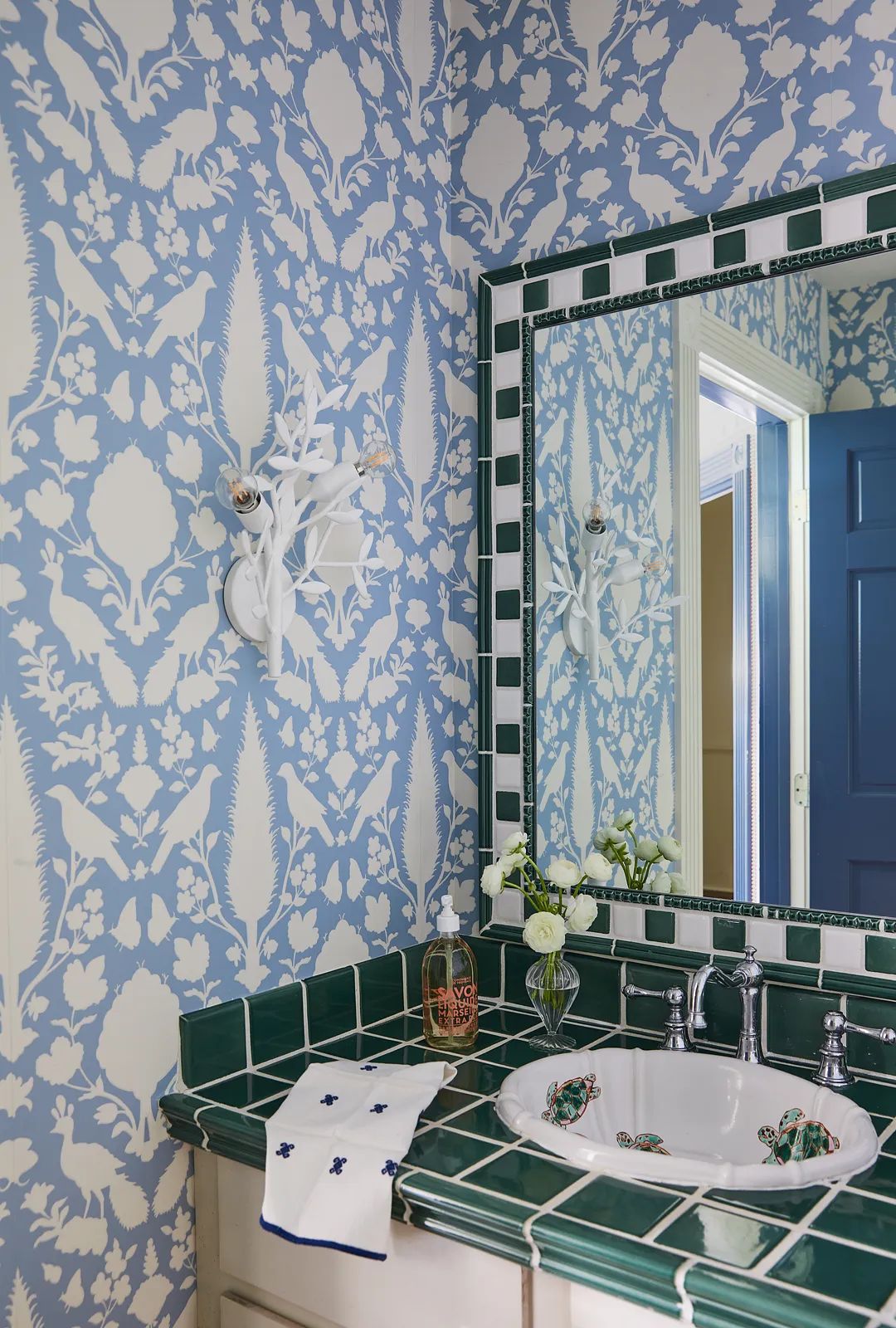 Bathroom with green tiled countertop, dual sinks, large mirror, and blue floral wallpaper.