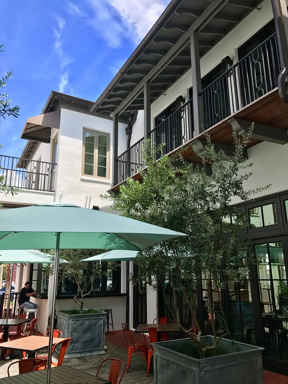 Exterior front view of a building with balconies, trees, umbrellas, and outdoor seating.
