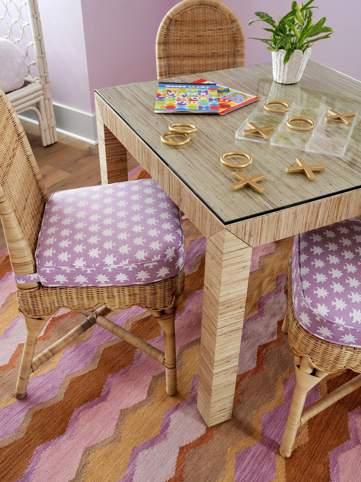 Dining room with glass table, wicker chairs, lavender cushions, area rug, and decorative plant.