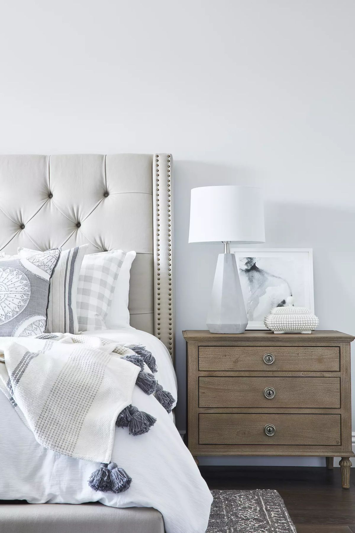 Bedroom with tufted headboard, white bedding, wooden nightstand, lamp, decorative boxes, and light area rug.