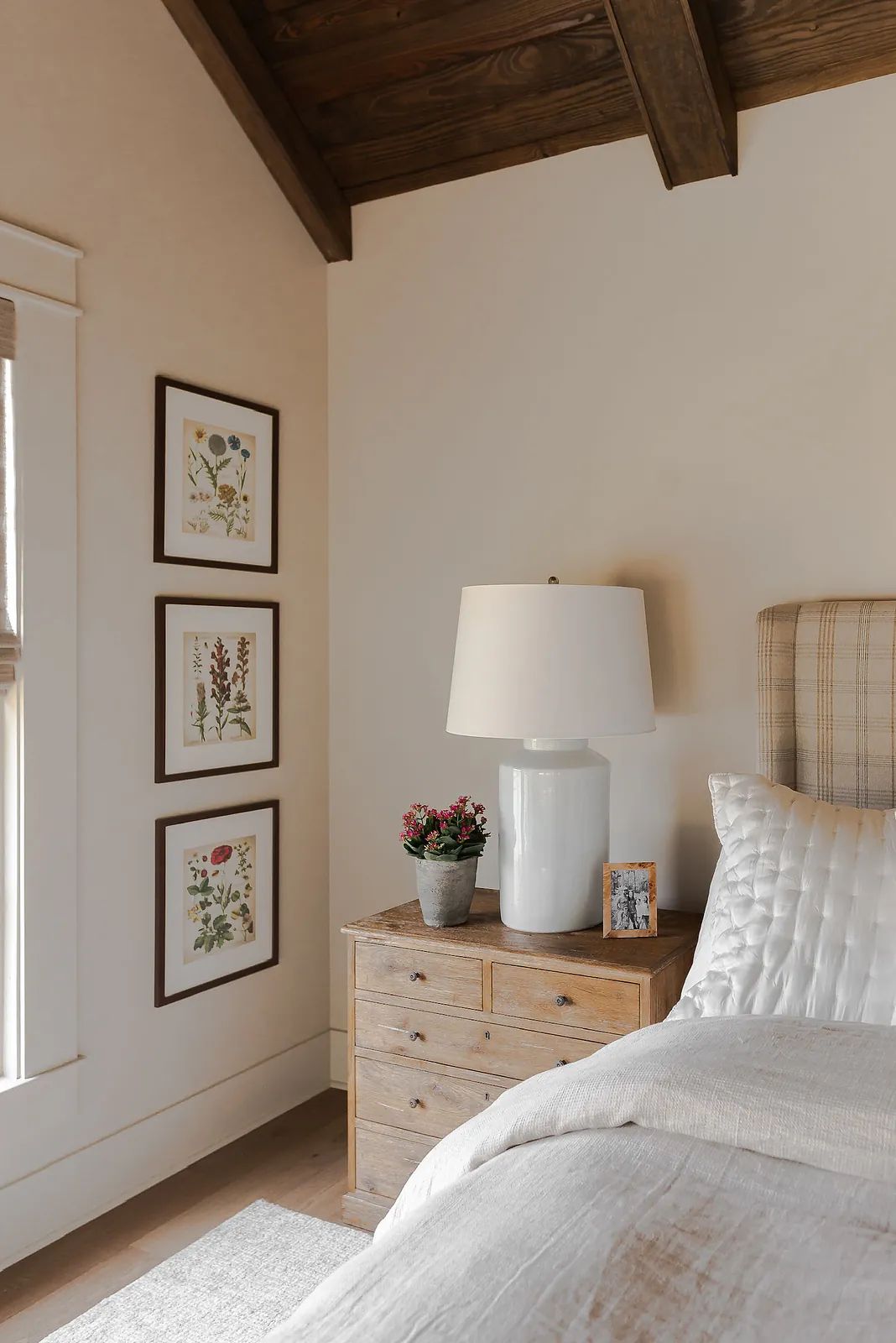 Bedroom with plaid headboard, white bedding, nightstand, lamp, artwork, and window with natural light