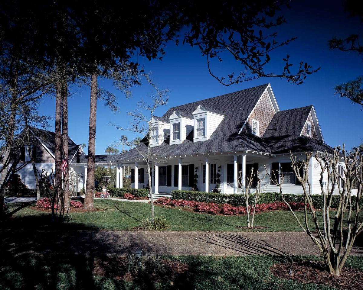 Front exterior of a two-story house with gabled roof, white siding, porch, flower beds, and trees