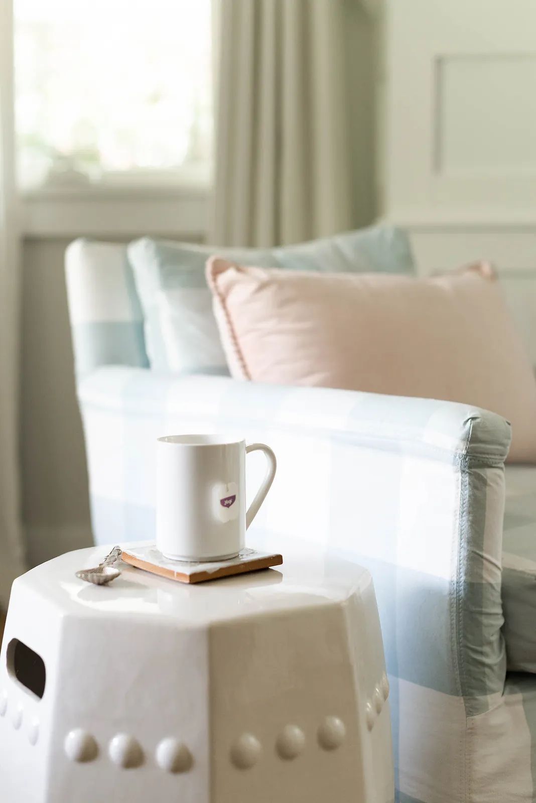 Living room with light blue checked armchair, pink cushion, white stool, and mug on a coaster near a window.