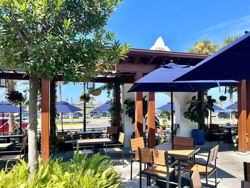 Outdoor patio with wooden tables, chairs, blue umbrellas, and surrounding potted plants