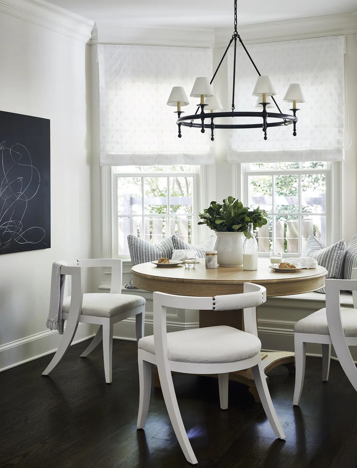Dining room with round wooden table, four white chairs, and large windows with shades.