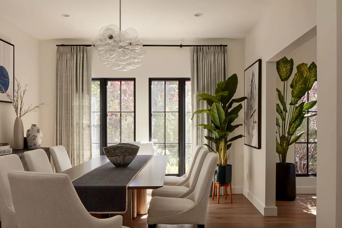 Dining room with wooden table, six chairs, centerpiece bowl, windows, and potted plants.