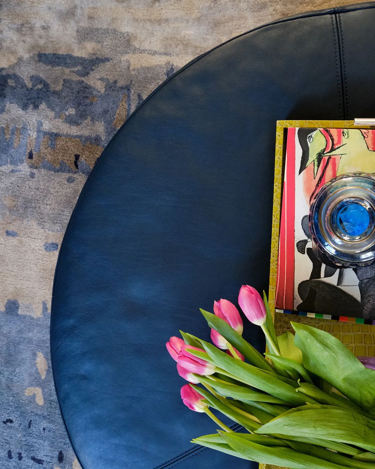 Close-up of round black leather table with art book, glass bowl, and pink tulips on an abstract rug.