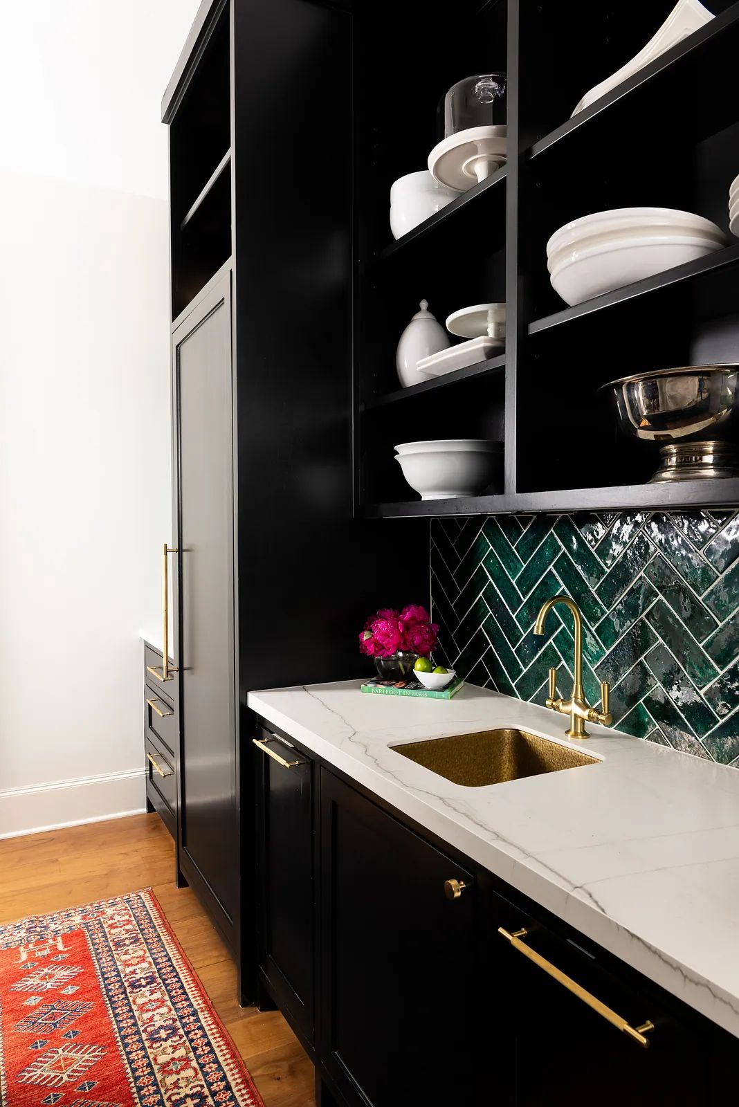 Kitchen with black cabinetry, white marble countertop, gold sink, emerald green backsplash, and patterned red rug