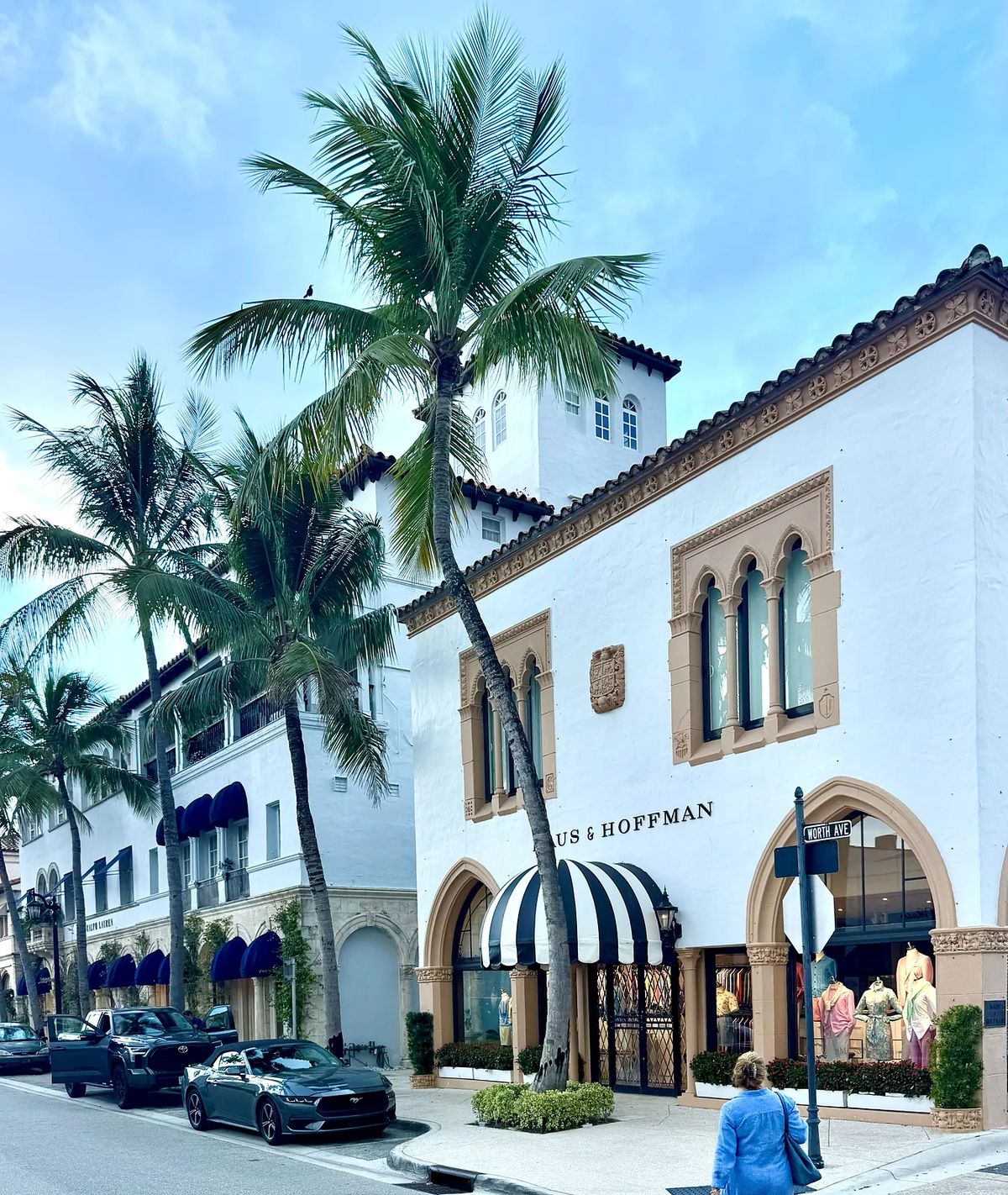 Exterior view of storefront with palm trees, arched windows, black and white awning, and parked cars.