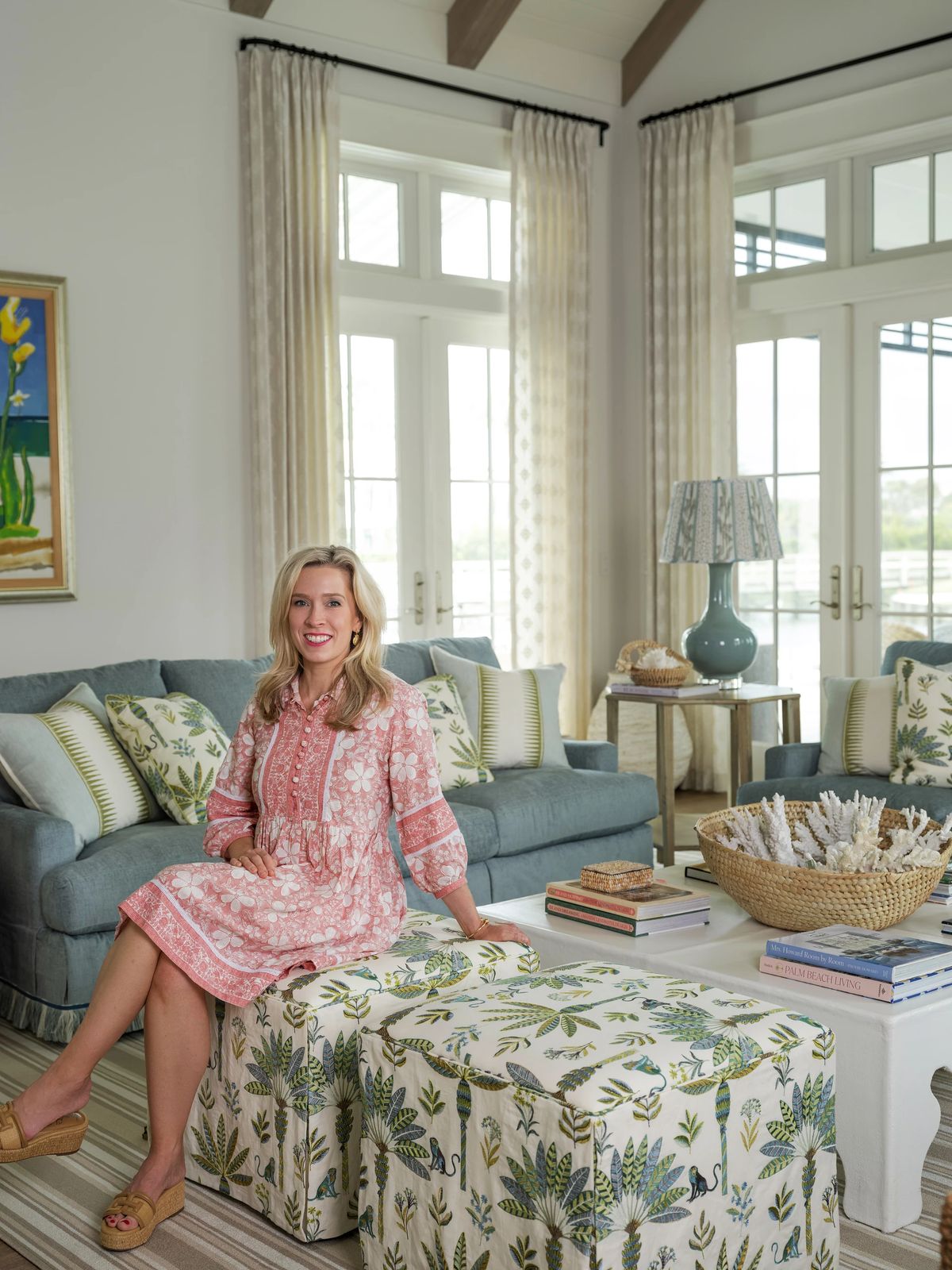 Living room with blue sofa, floral stools, coffee table with books, large windows, and patterned lamp.