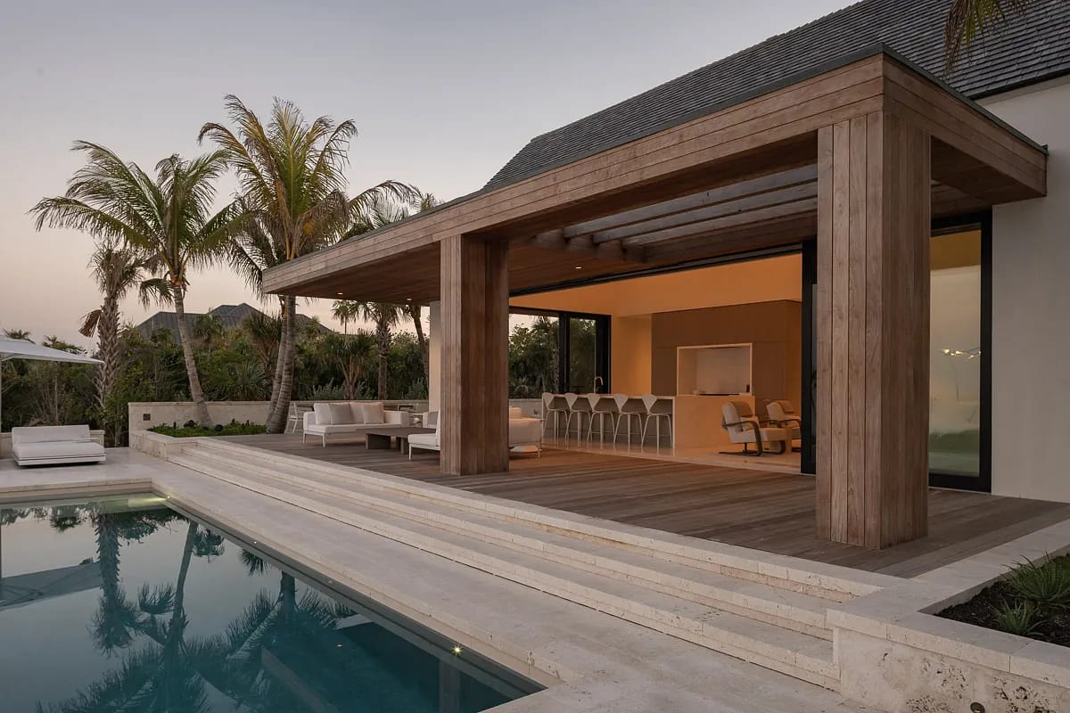 Outdoor patio with wooden pergola, sectional sofa, dining table, chairs, and swimming pool with palm trees.