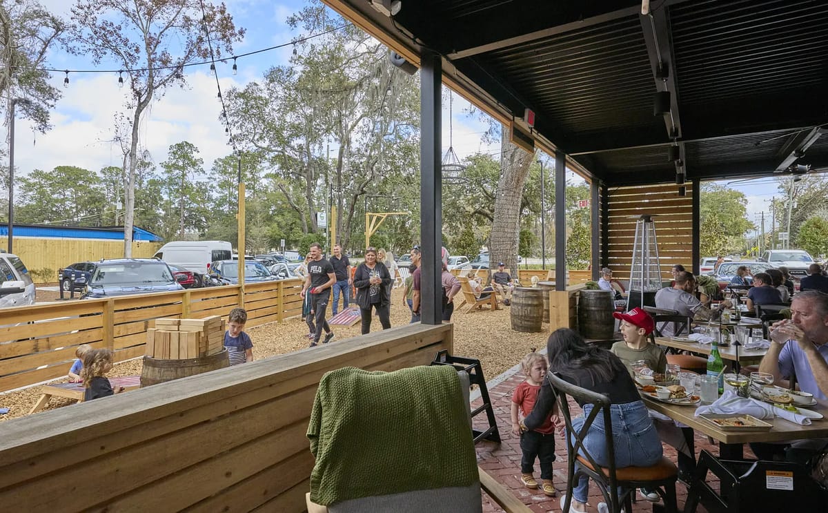 Outdoor patio with wooden benches, metal chairs, tables, and dining guests, surrounded by trees and fencing.