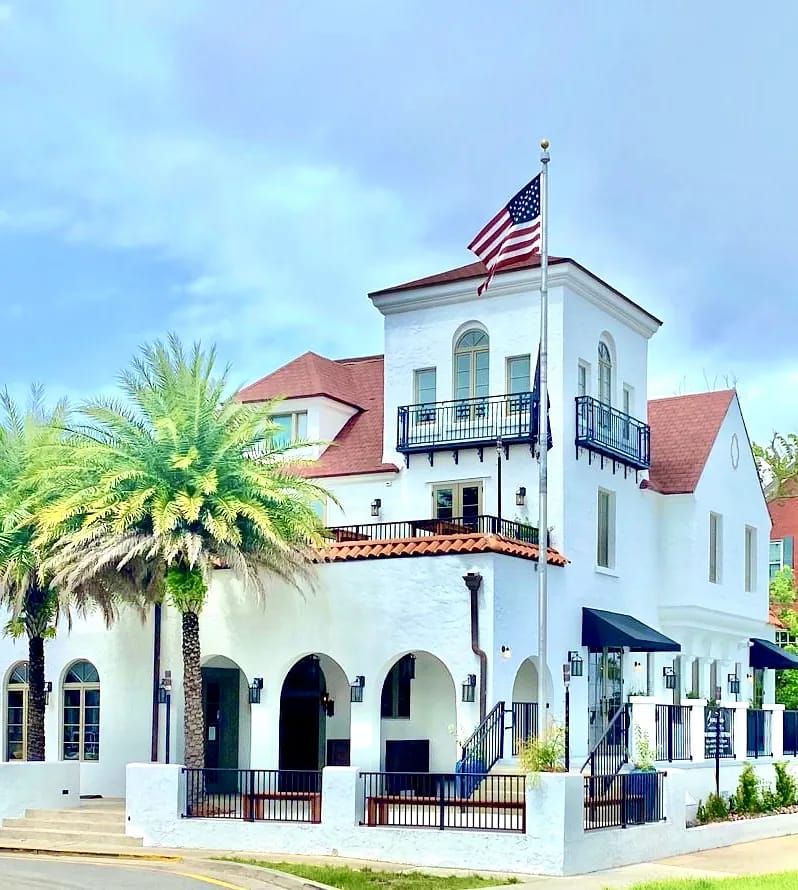 Front view of white building with tile roof, balcony, palm trees, and American flag.