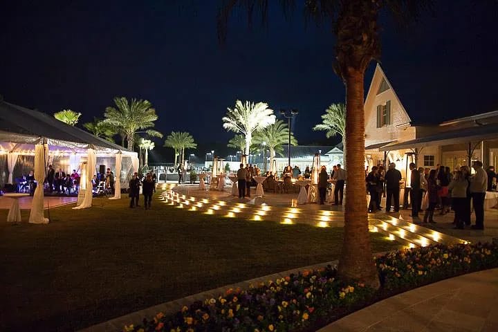 Outdoor garden space with palm trees, tents, tables, guests, and illuminated pathways at night.