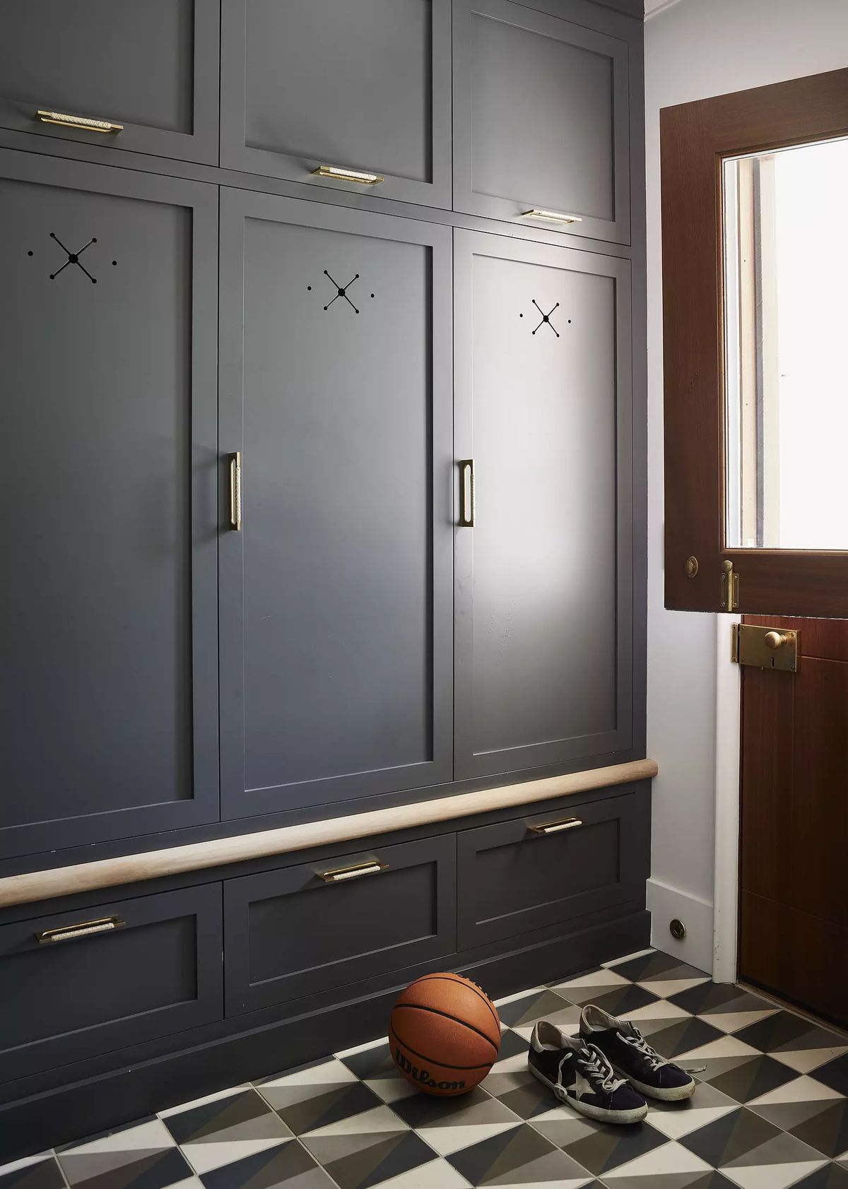 Mudroom with dark cabinetry, wooden bench, basketball, shoes, and patterned tile floor with natural light.