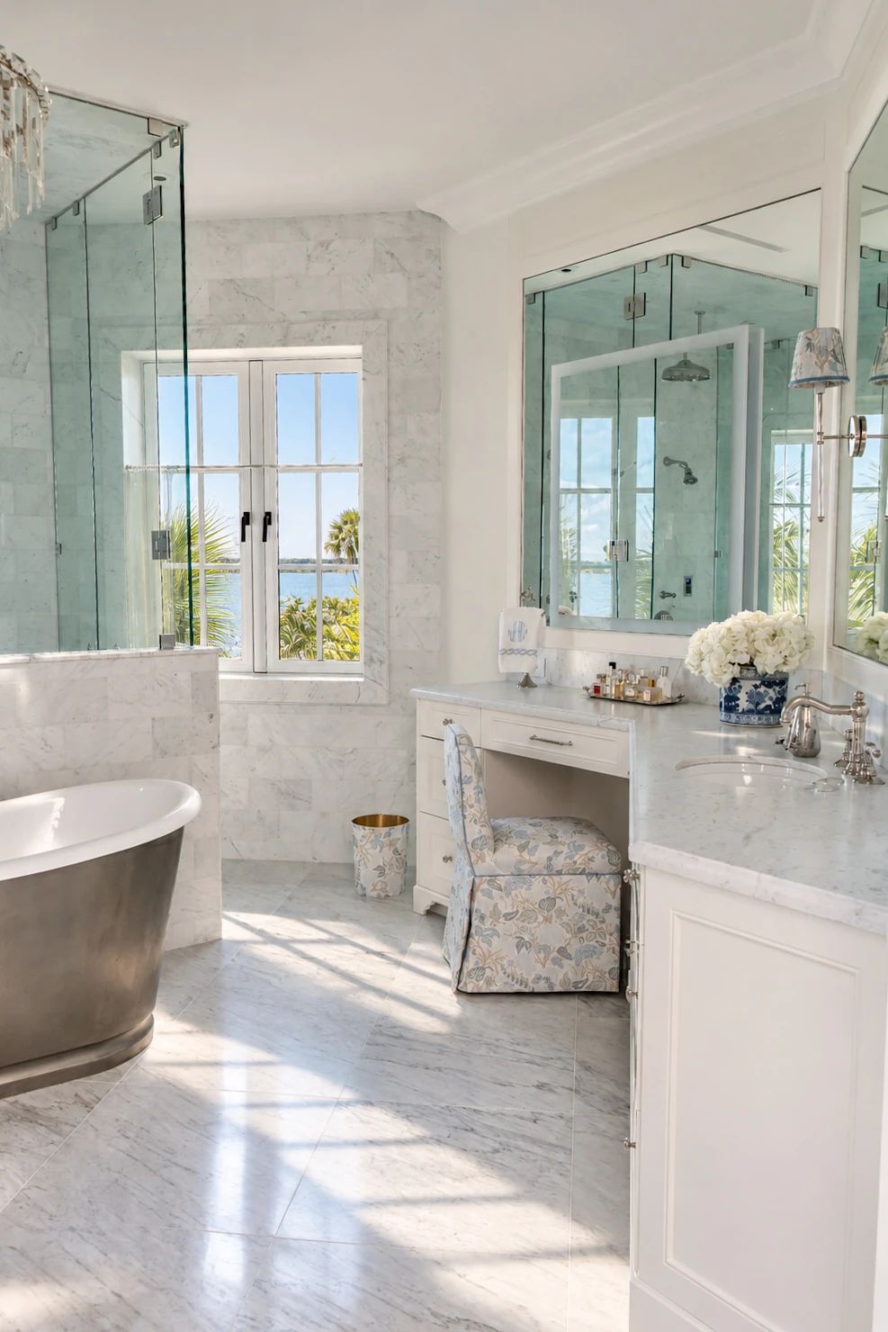 Bathroom with freestanding tub, dual vanities, glass shower, marble surfaces, and floral-patterned chair.