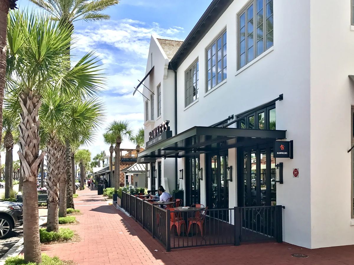 Exterior front view of a building with large windows, patio seating, and palm trees along a red brick pathway.