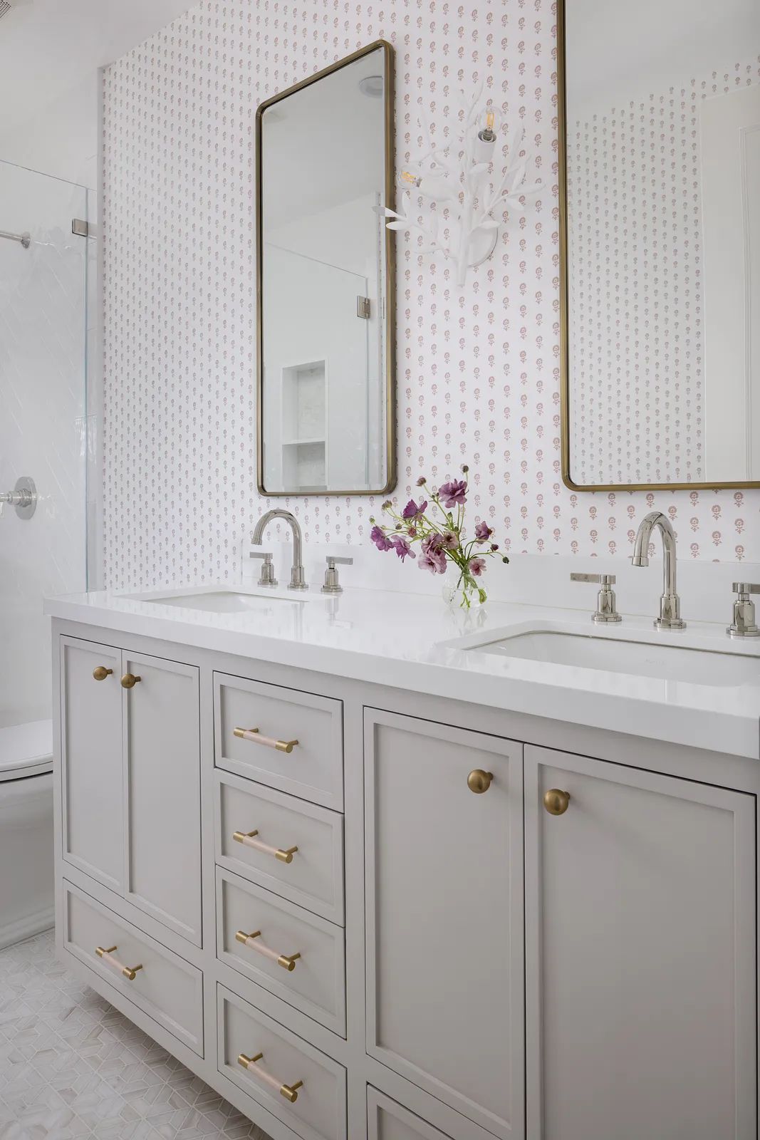 Bathroom with gray double vanity, white countertop, patterned wallpaper, and glass shower enclosure.