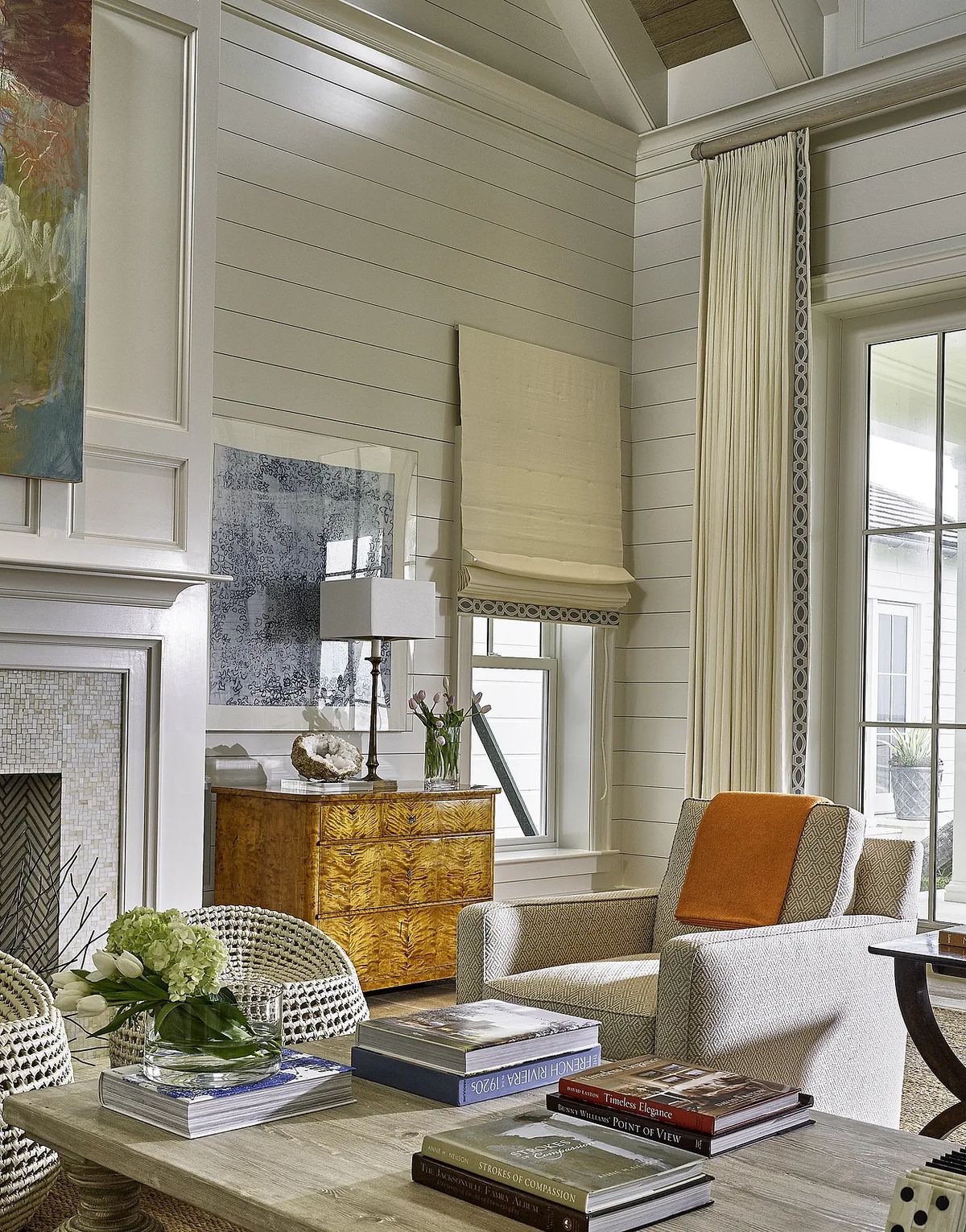 Living room with beige armchair, wooden side table, decorative books, large window, and fireplace with mosaic front.
