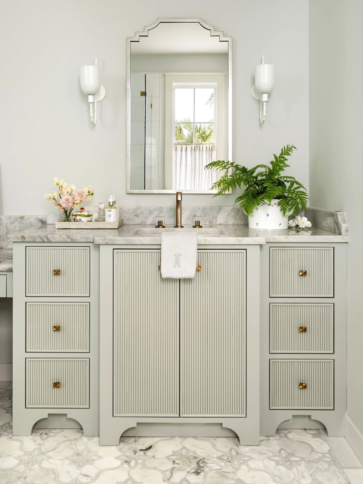 Bathroom with gray vanity, white countertop, scalloped mirror, wall sconces, and marbled floor tiles.