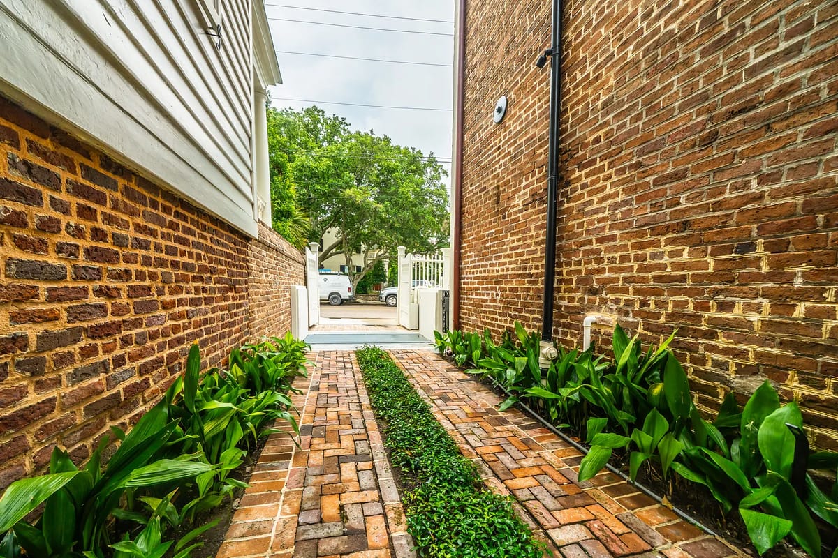 Narrow garden path with green plants and yellow brick pavers, leading to a white gate.