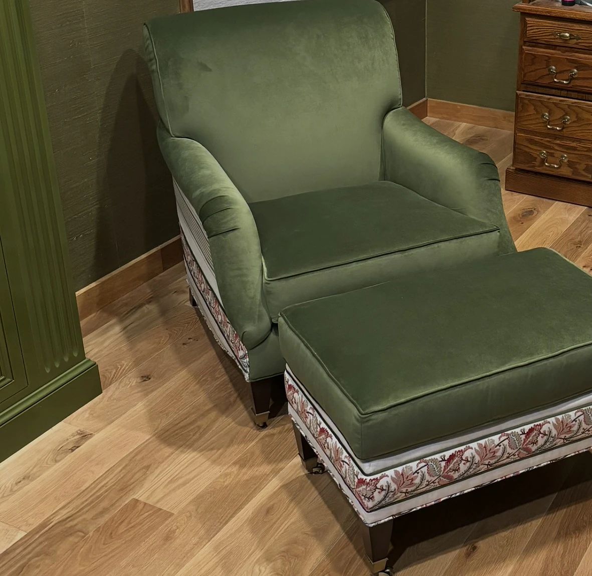 Seating area with green armchair, matching ottoman, and wooden nightstand on hardwood flooring.