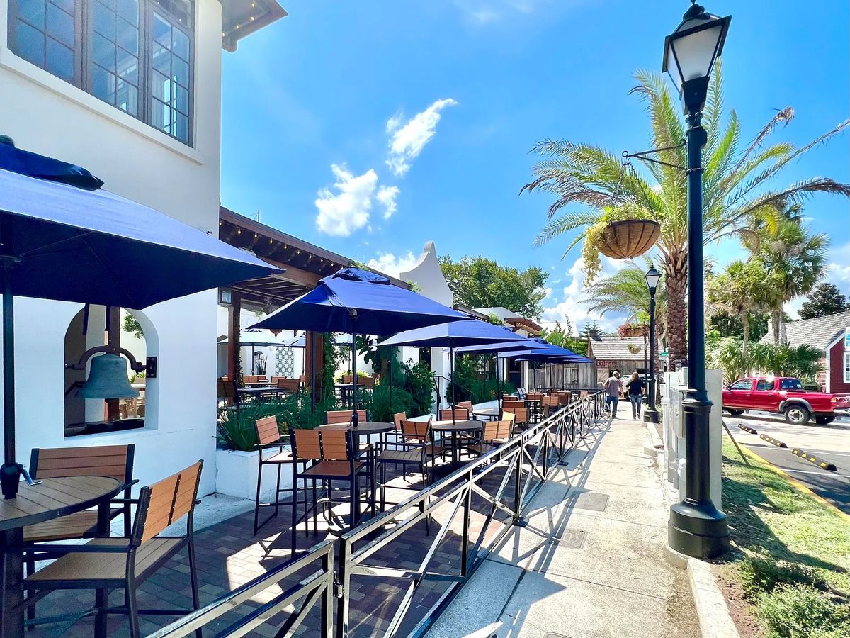 Outdoor patio with wooden tables, chairs, blue umbrellas, and landscaped foliage.