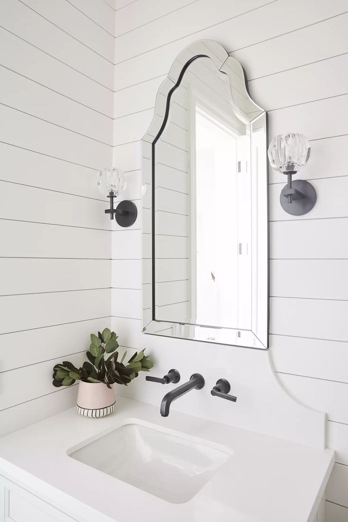 Bathroom with white vanity, ornate mirror, black faucets, and shiplap paneling with a plant on the vanity.