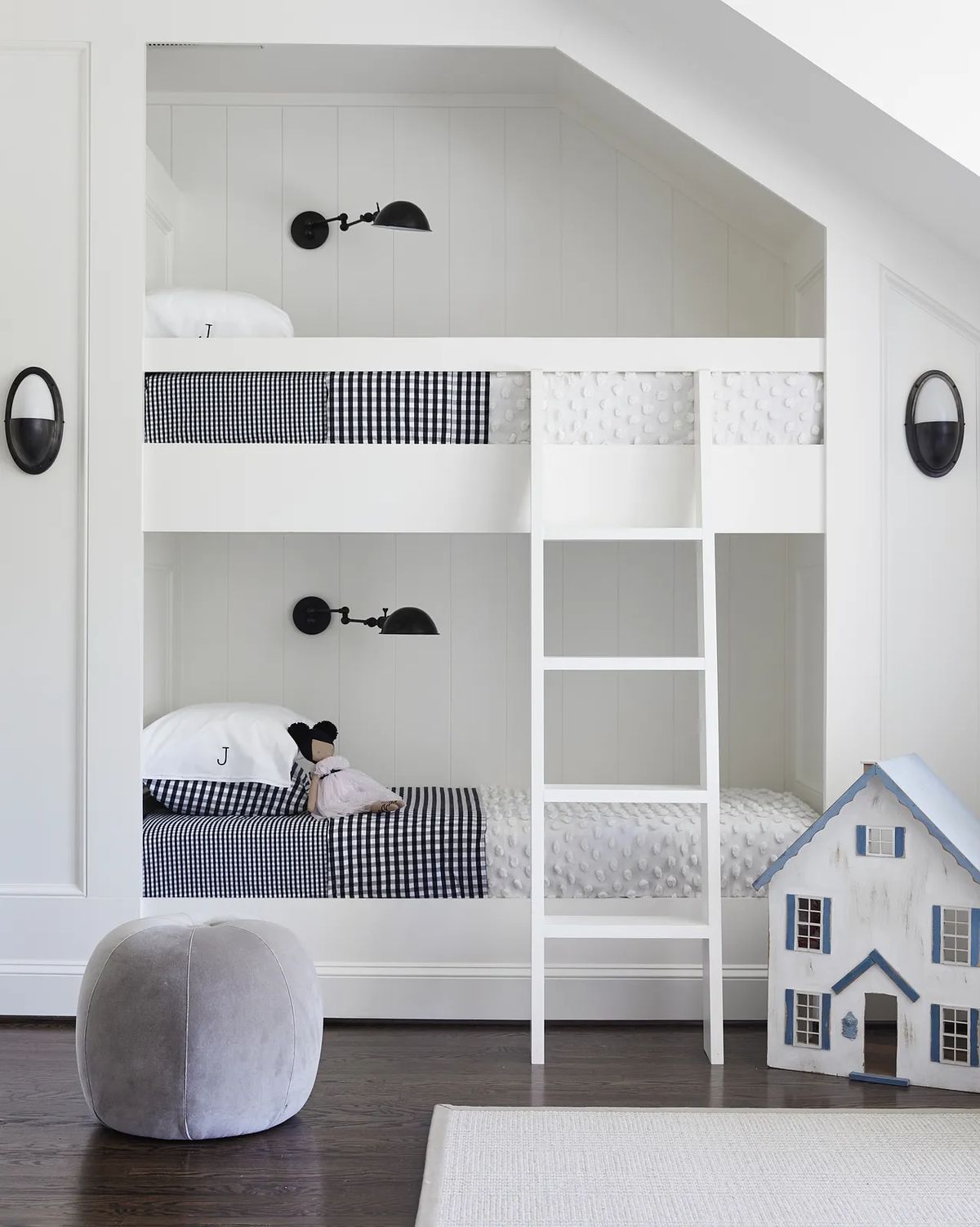 Bedroom with white bunk bed, checkered bedding, gray pouf, and dollhouse on a paneled wall.