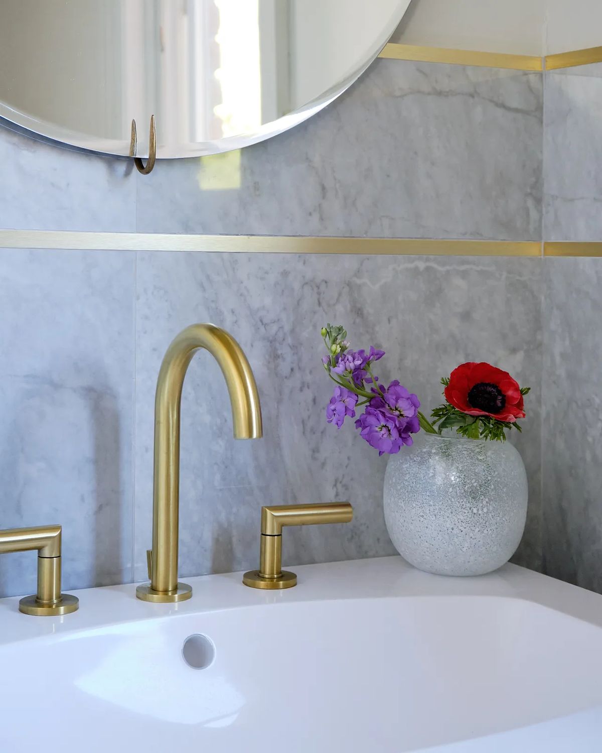 Bathroom with freestanding tub, gold faucets, round mirror, marbled wall, and a vase of flowers
