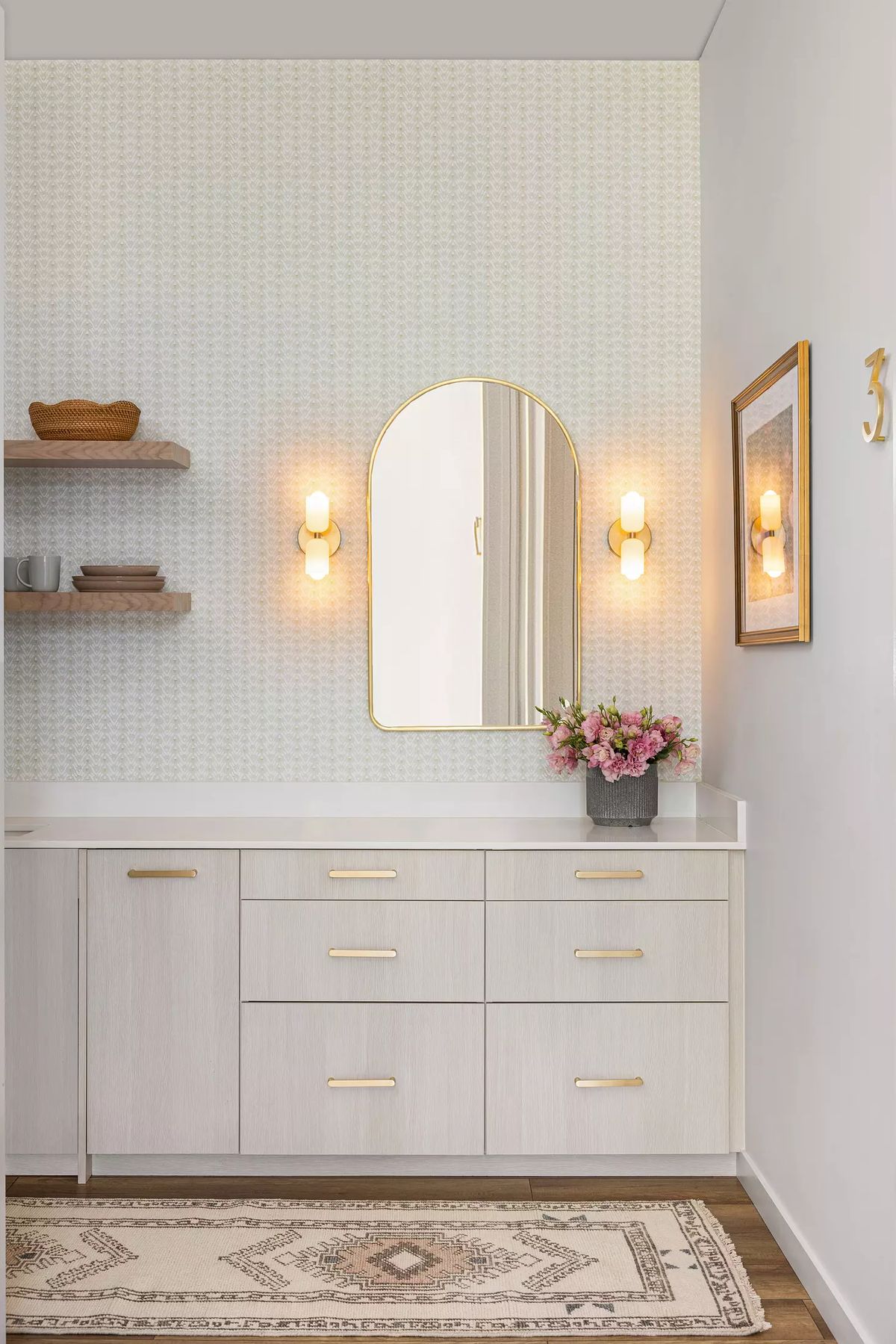 Powder room with light vanity, gold-framed mirror, wall sconces, patterned wallpaper, and a vase of flowers