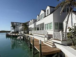 Waterfront house with wooden deck, gabled roofs, large windows, palm trees, and moored boats.