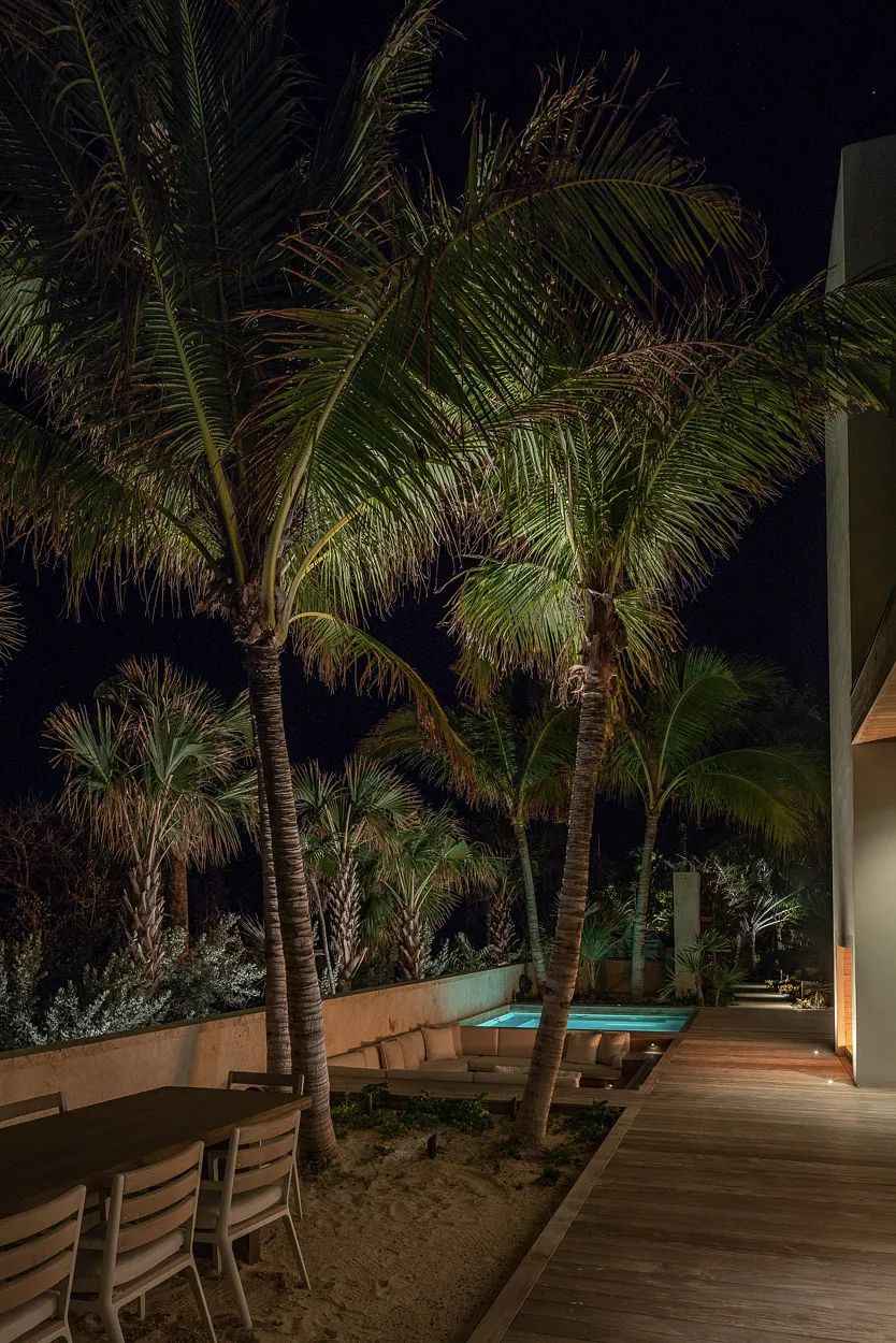 Outdoor patio with wooden dining table, chairs, palm trees, and swimming pool visible at night