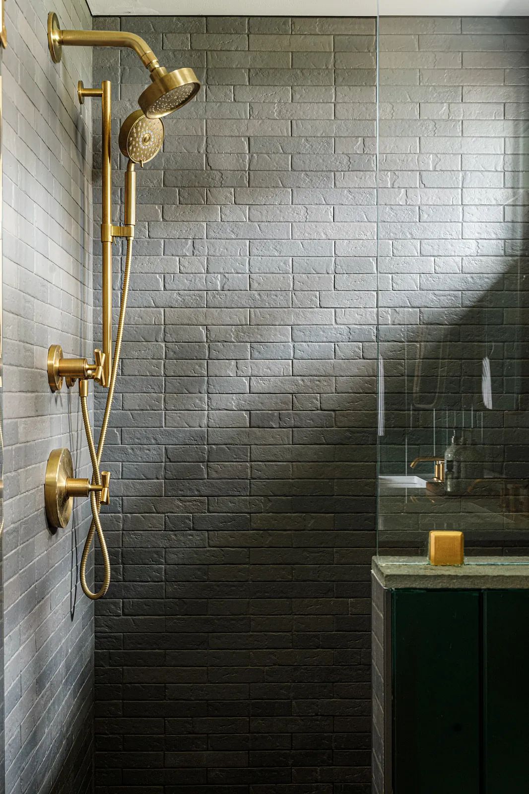 Bathroom with walk-in shower, gold fixtures, gray brick tiles, and dark green vanity