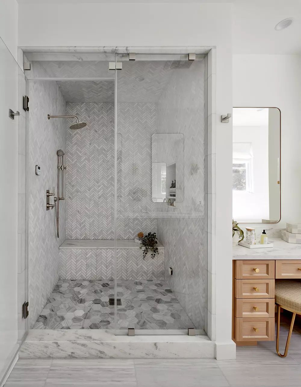 Bathroom with glass shower enclosure, herringbone tile, vanity, large mirror, and plants on the counter.