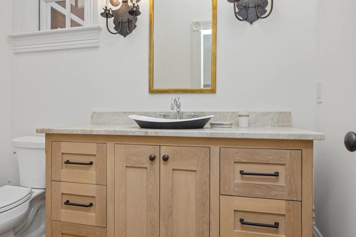 Powder room with wood vanity, beige countertop, black vessel sink, round gold-framed mirror, and two sconces.
