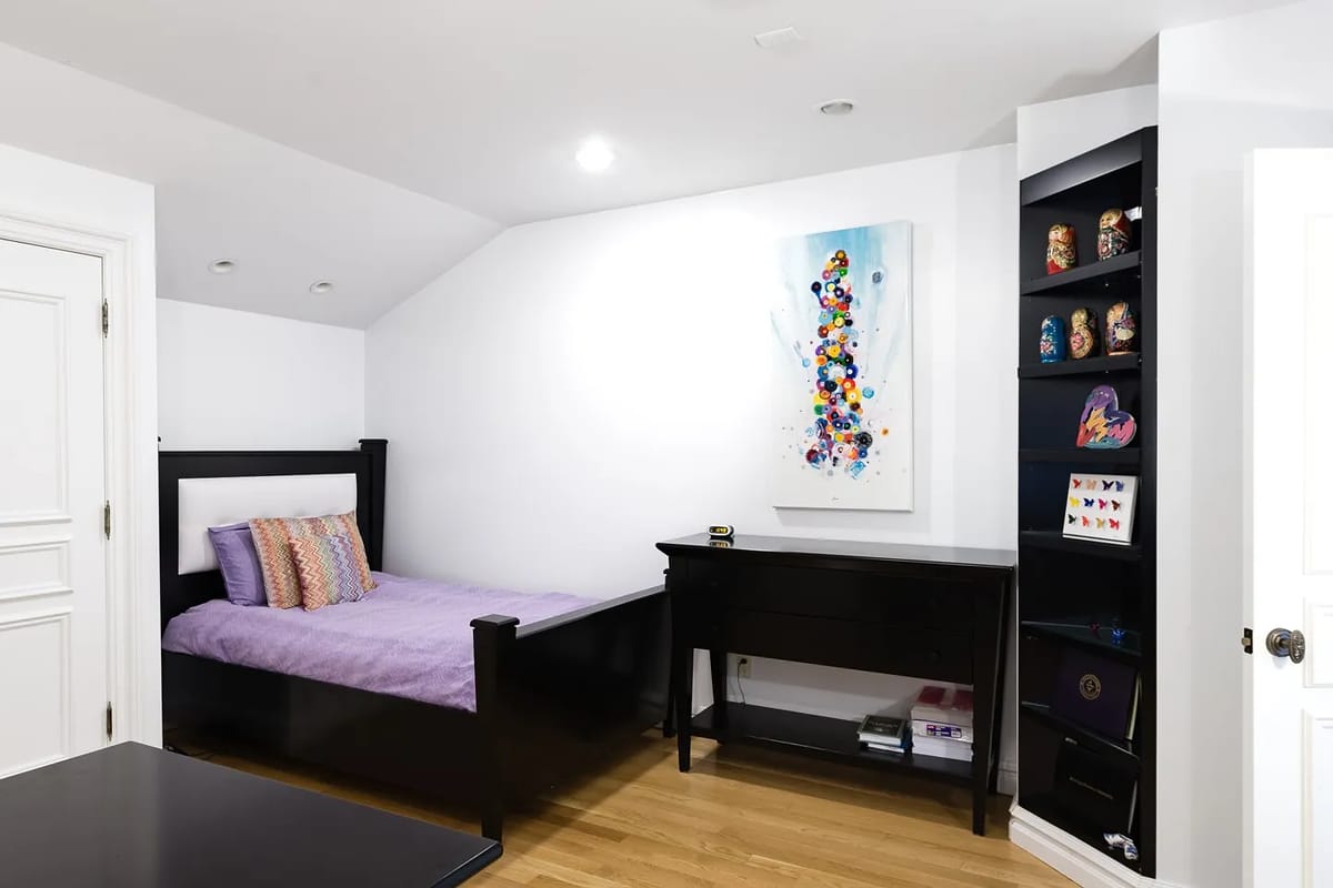 Bedroom with black bed, purple blanket, desk, wall-mounted shelf, and colorful painting on sloped wall.