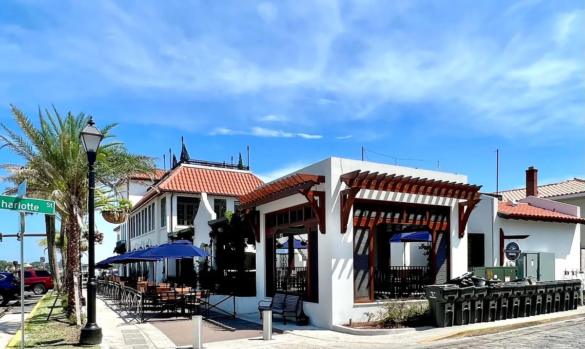 Exterior view of a restaurant with white facade, red tiled roof, and outdoor seating with blue umbrellas.