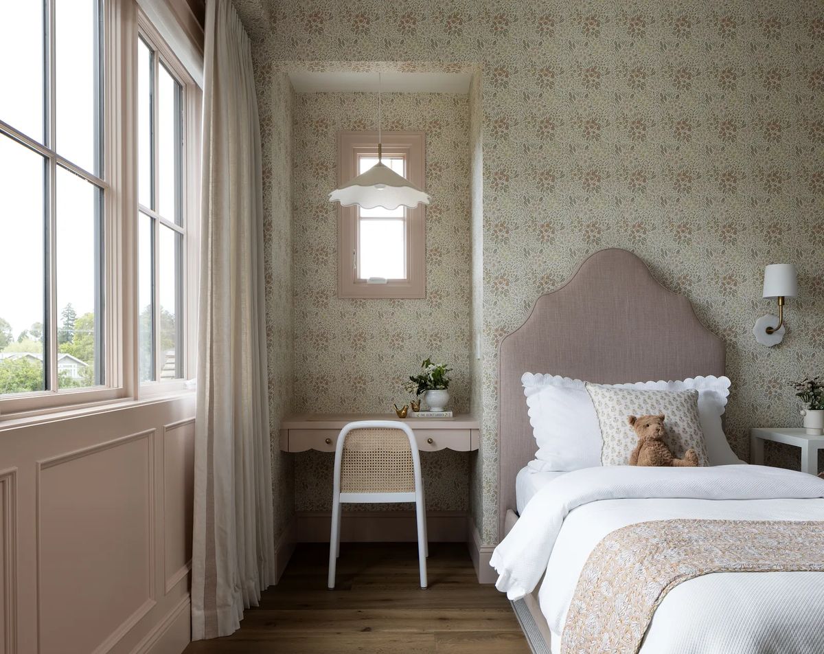 Bedroom with pink upholstered headboard, white bedding, floral wallpaper, desk with rattan chair, and teddy bear.